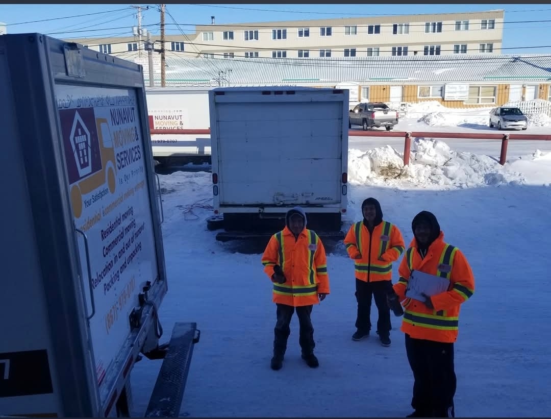 Three NMS team members in orange safety vests standing beside branded moving trucks in Iqaluit